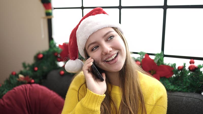 νεαρή ξανθιά γυναίκα που μιλάει στο smartphone καθισμένη στον καναπέ με χριστουγεννιάτικη διακόσμηση στο σπίτι στοκ εικόνα