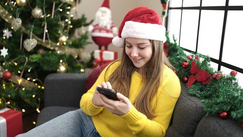 νεαρή ξανθιά γυναίκα που χρησιμοποιεί smartphone καθισμένη στον καναπέ δίπλα στο χριστουγεννιάτικο δέντρο στο σπίτι στοκ εικόνες