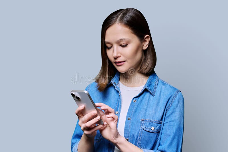 Young woman using smartphone in gray background. Serious 20s female looking at screen, texting. Mobile Internet applications apps, technologies for work education communication shopping healthcare. Young woman using smartphone in gray background. Serious 20s female looking at screen, texting. Mobile Internet applications apps, technologies for work education communication shopping healthcare