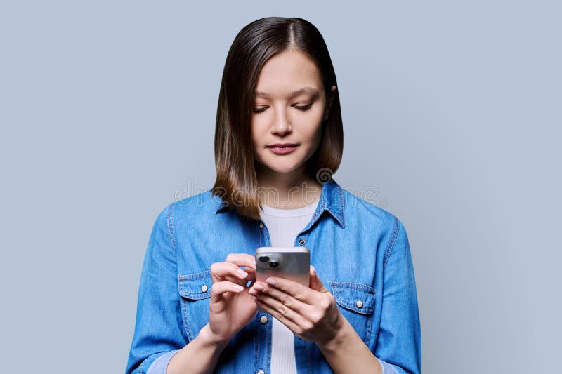 Young woman using smartphone in gray background. Serious 20s female looking at screen, texting. Mobile Internet applications apps, technologies for work education communication shopping healthcare. Young woman using smartphone in gray background. Serious 20s female looking at screen, texting. Mobile Internet applications apps, technologies for work education communication shopping healthcare