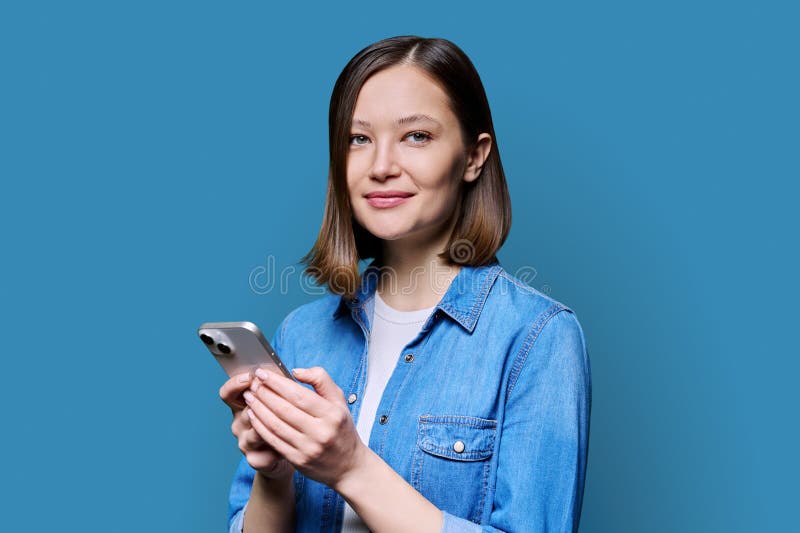 Young happy woman using smartphone in blue background. Smiling 20s female looking at camera texting. Mobile Internet apps applications technologies for work education communication shopping healthcare. Young happy woman using smartphone in blue background. Smiling 20s female looking at camera texting. Mobile Internet apps applications technologies for work education communication shopping healthcare