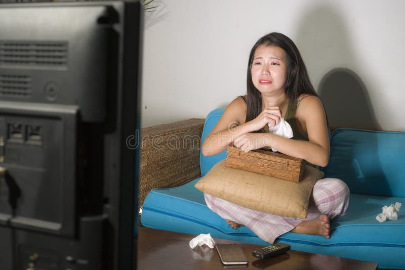 Funny lifestyle portrait of young cute and emotional Asian Chinese woman watching korean drama show on TV sitting at home living room couch crying wiping tears with tissue paper. Funny lifestyle portrait of young cute and emotional Asian Chinese woman watching korean drama show on TV sitting at home living room couch crying wiping tears with tissue paper
