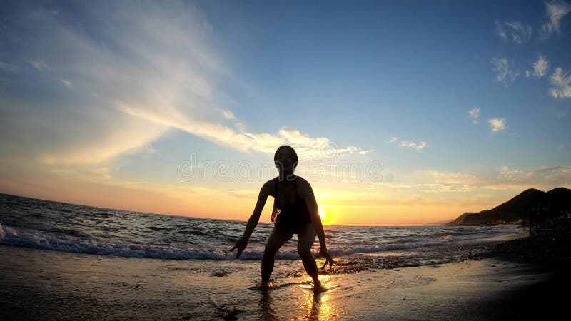 νεαρή χαρούμενη γυναίκα πηδάει στο χρυσό ηλιοβασίλεμα στην αμμώδη παραλία στα κύματα της θάλασσας