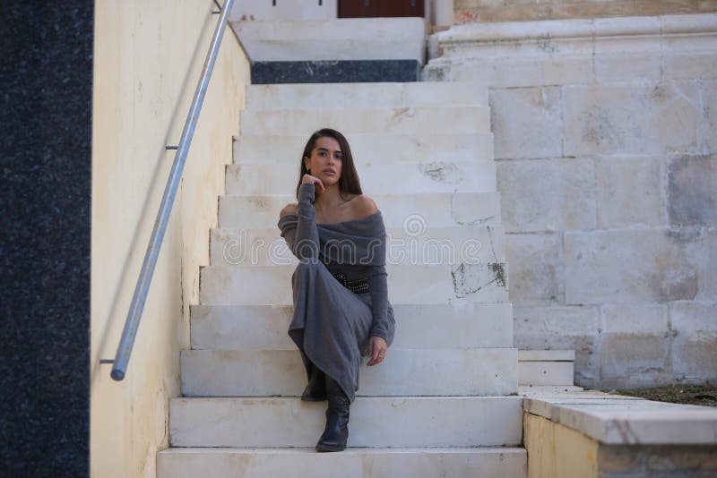 Νεαρή όμορφη μελαχρινή γυναίκα με γκρι φόρεμα και μπότες που ποζάρουν κοιτώντας την κάμερα να κάθεται στις σκάλες. Έννοια στοκ εικόνα