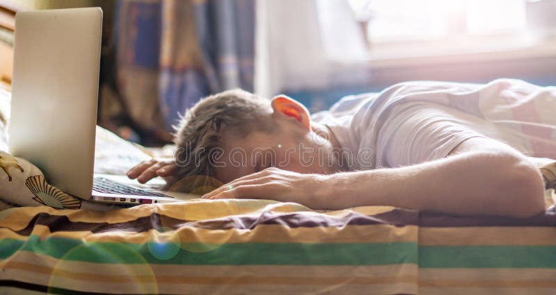 Νεαρό άτομο που βρίσκεται και που κοιμάται στο κρεβάτι στο ηλιόλουστο πρωί μετά από να εργαστεί στο lap-top στη νύχτα φ στοκ φωτογραφίες