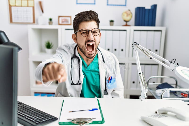 Young man with beard wearing doctor uniform and stethoscope at the clinic pointing displeased and frustrated to the camera, angry and furious with you. Young man with beard wearing doctor uniform and stethoscope at the clinic pointing displeased and frustrated to the camera, angry and furious with you