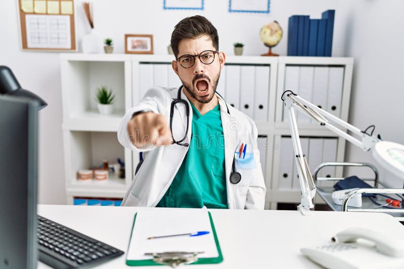 Young man with beard wearing doctor uniform and stethoscope at the clinic pointing displeased and frustrated to the camera, angry and furious with you. Young man with beard wearing doctor uniform and stethoscope at the clinic pointing displeased and frustrated to the camera, angry and furious with you