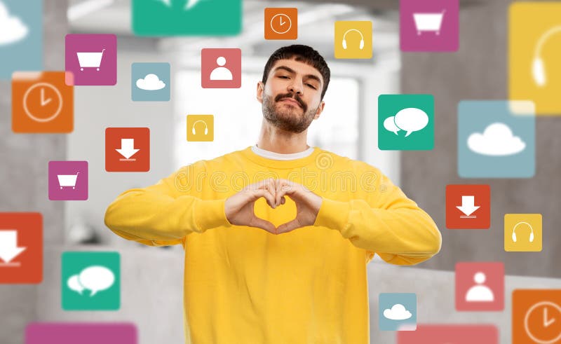 love, valentine&#x27;s day and people concept - young man in yellow sweatshirt girl making hand heart gesture over mobile app icons and office background. love, valentine&#x27;s day and people concept - young man in yellow sweatshirt girl making hand heart gesture over mobile app icons and office background