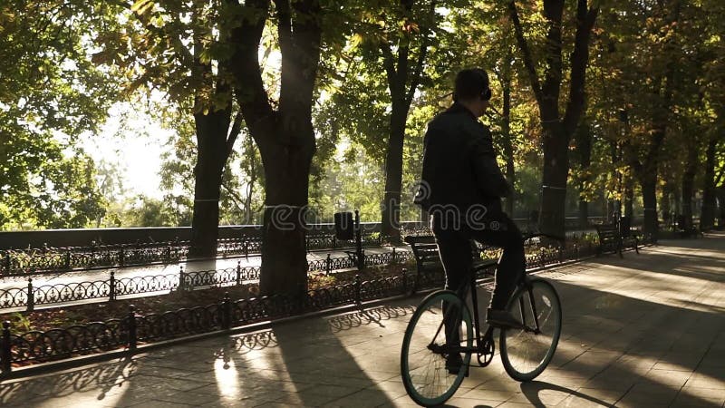 Νεαρός άνδρας με τα ακουστικά που οδηγούν το ποδήλατο στο πάρκο στο μαύρο ποδήλατο οδοιπορίας Οδήγηση και χορός ακούοντας