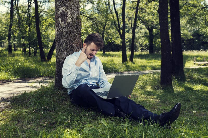 Νεαρός άνδρας που εργάζεται στο lap-top του σε μια δημόσια συνεδρίαση πάρκων στο έδαφος και που κάνει τη χειρονομία επιτυχίας στοκ εικόνες Νεαρός άνδρας που εργάζεται στο lap-top του σε μια δημόσια συνεδρίαση πάρκων στο έδαφος και που κάνει τη χειρονομία επιτυχίας στοκ εικόνες