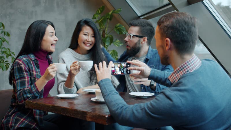 νεαρός άνδρας που βγάζει φωτογραφίες ευτυχισμένων φίλων που ποζάρουν με φλιτζάνια καφέ στο σύγχρονο καφέ