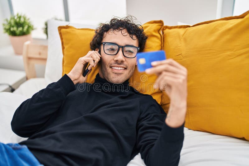 νεαρός ισπανός άνδρας μιλάει στο smartphone κρατώντας πιστωτική κάρτα ξαπλωμένη στο κρεβάτι στοκ φωτογραφία με δικαίωμα ελεύθερης χρήσης