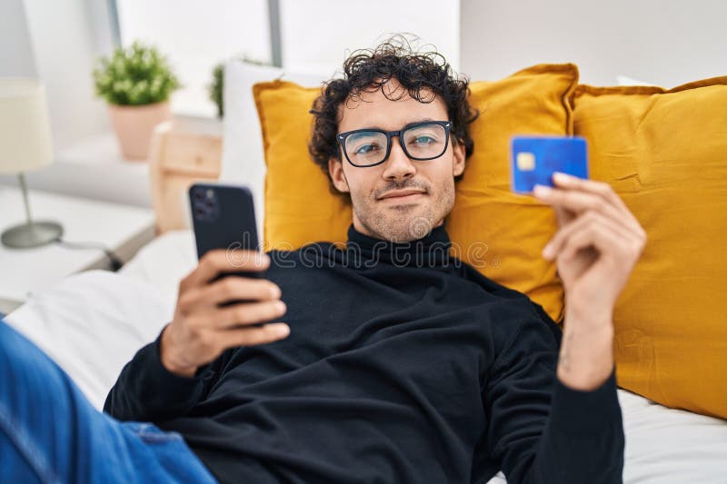νεαρός ισπανός που χρησιμοποιεί smartphone και πιστωτική κάρτα ξαπλωμένη στο κρεβάτι στοκ φωτογραφίες