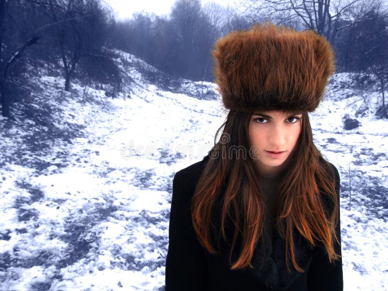 A view of young woman with bright reddish hair and wearing a tall Cossack fur hat, in the forest on a gray winter day. A view of young woman with bright reddish hair and wearing a tall Cossack fur hat, in the forest on a gray winter day