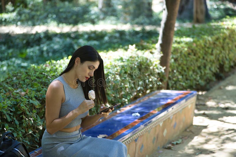 South American woman, young, pretty, brunette in gray top eating an ice cream while checking her mobile phone, sitting on a bench. Concept beauty, food, summer, ice cream, heat. South American woman, young, pretty, brunette in gray top eating an ice cream while checking her mobile phone, sitting on a bench. Concept beauty, food, summer, ice cream, heat