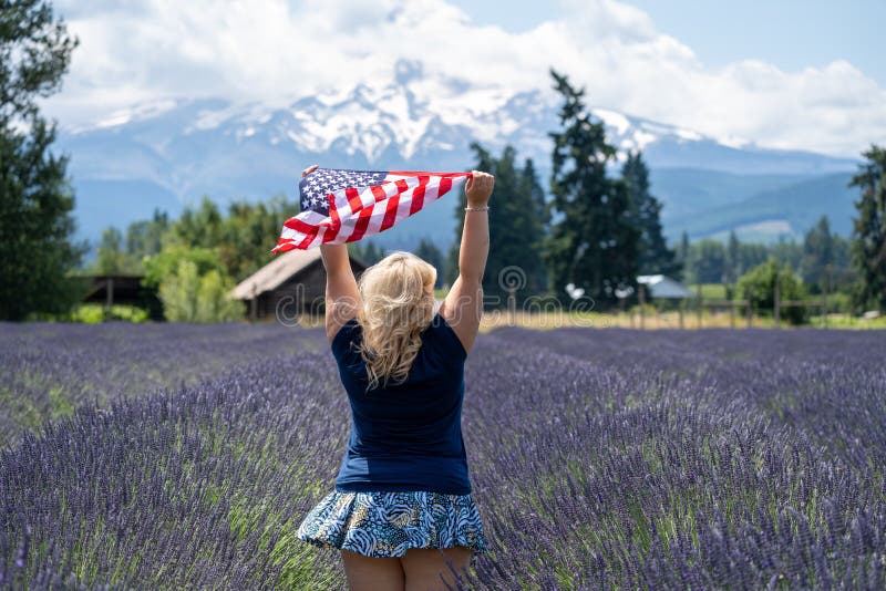 Ξανθή γυναίκα που στέκεται σε έναν τομέα Lavender κοντά στην ΑΜ Η κουκούλα κρατά ένα αμερικανικό Fiag όπως φυσά στον αέρα Έννοια στοκ εικόνες Ξανθή γυναίκα που στέκεται σε έναν τομέα Lavender κοντά στην ΑΜ Η κουκούλα κρατά ένα αμερικανικό Fiag όπως φυσά στον αέρα Έννοια στοκ εικόνες