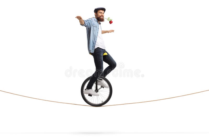 Ο γενειοφόρος νεαρός άνδρας που οδηγά ένα unicycle σε ένα σχοινί και που κρατά ένα κόκκινο αυξήθηκε με το στόμα του
