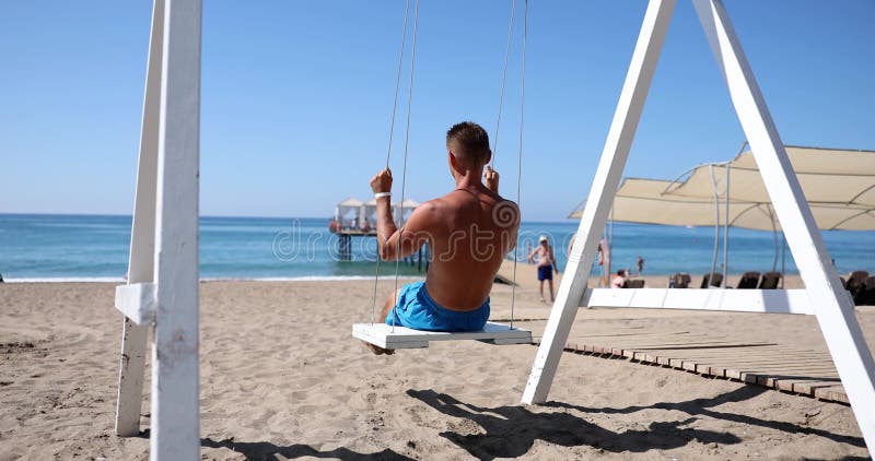 Ο γυμνοσάλιαγκας άνδρας με μπλε σορτσάκια απολαμβάνει την ταλάντευση στην ηλιόλουστη παραλία φιλμ μικρού μήκους