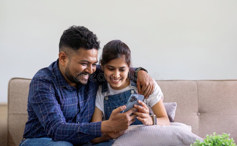 The triumph of a young Indian couple happily celebrates reading unexpected good news on their smartphone. young Indian couple together on the sofa in the living room. The triumph of a young Indian couple happily celebrates reading unexpected good news on their smartphone. young Indian couple together on the sofa in the living room.