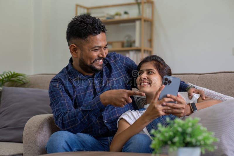 The triumph of a young Indian couple happily celebrates reading unexpected good news on their smartphone. young Indian couple together on the sofa in the living room. The triumph of a young Indian couple happily celebrates reading unexpected good news on their smartphone. young Indian couple together on the sofa in the living room.