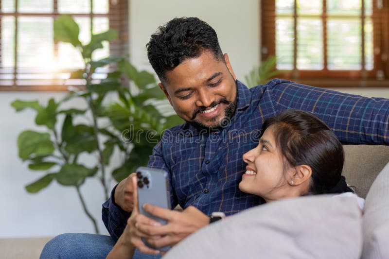 The triumph of a young Indian couple happily celebrates reading unexpected good news on their smartphone. young Indian couple together on the sofa in the living room. The triumph of a young Indian couple happily celebrates reading unexpected good news on their smartphone. young Indian couple together on the sofa in the living room.