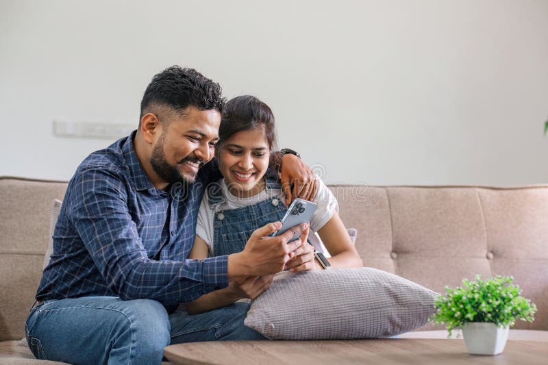 The triumph of a young Indian couple happily celebrates reading unexpected good news on their smartphone. young Indian couple together on the sofa in the living room. The triumph of a young Indian couple happily celebrates reading unexpected good news on their smartphone. young Indian couple together on the sofa in the living room.