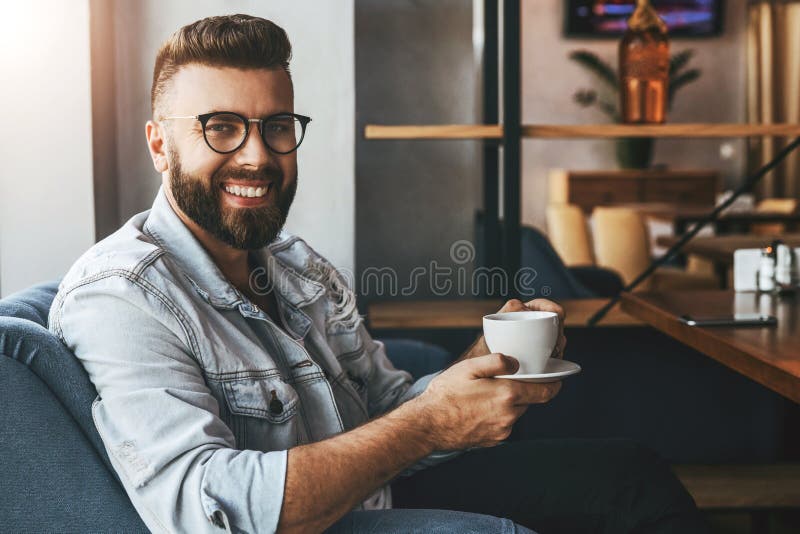 Ο νέος ελκυστικός γενειοφόρος επιχειρηματίας στα καθιερώνοντα τη μόδα γυαλιά κάθεται στον καφέ, πίνει τον καφέ κατά τη διάρκεια τ στοκ φωτογραφία Ο νέος ελκυστικός γενειοφόρος επιχειρηματίας στα καθιερώνοντα τη μόδα γυαλιά κάθεται στον καφέ, πίνει τον καφέ κατά τη διάρκεια τ στοκ φωτογραφία