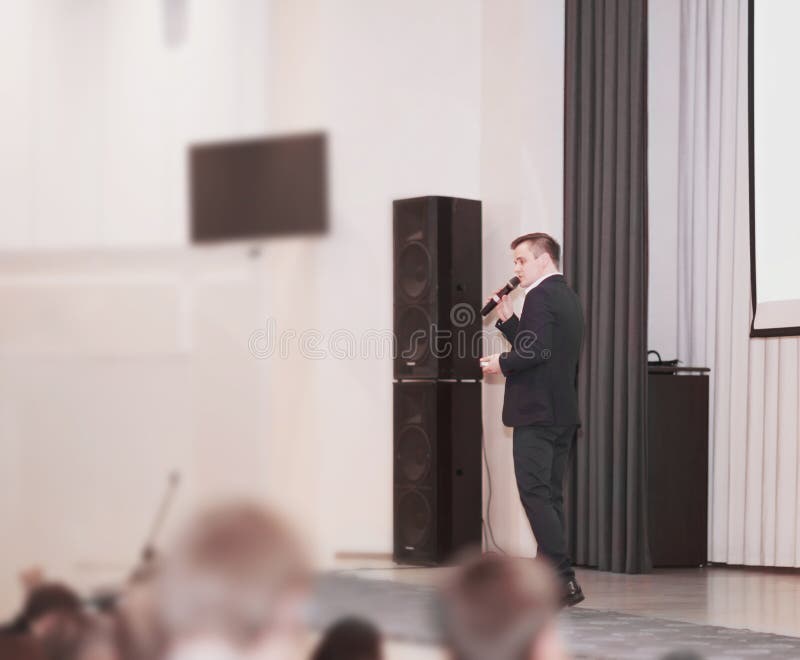 Ο ομιλητής διευθύνει την επιχείρηση της διάσκεψης που στέκεται μπροστά από μια μεγάλη άσπρη οθόνη στο στάδιο στη διάσκεψη στοκ εικόνα Ο ομιλητής διευθύνει την επιχείρηση της διάσκεψης που στέκεται μπροστά από μια μεγάλη άσπρη οθόνη στο στάδιο στη διάσκεψη στοκ εικόνα