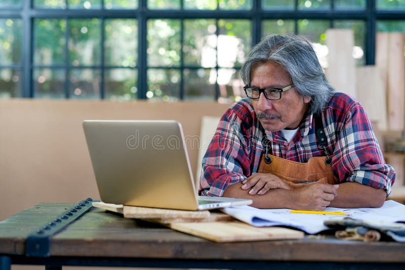 Ο παλιός Ασιάτης τεχνίτης κοιτάζει την οθόνη του φορητού υπολογιστή και σκέφτεται τη διαδικασία του για εργασία και κάθεται κοντά στοκ εικόνες
