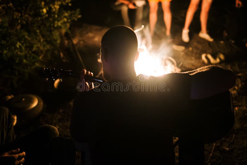 Ο χίπστερ παίζει με την ακουστική κιθάρα και τραγουδάει σε μεγάλη φωτιά στην κατασκήνωση, κάνοντας κρύο με φίλους στην Ομάδα Δασώ στοκ φωτογραφίες