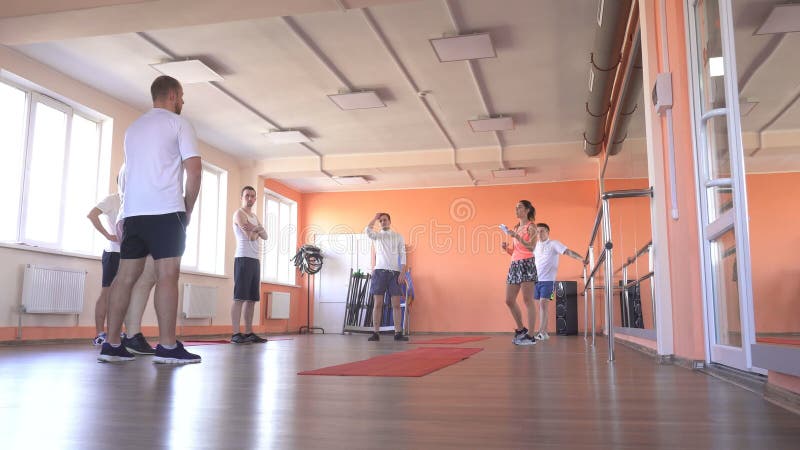 Ο όμορφος καυκάσιος εκπαιδευτής κοριτσιών στη γυμναστική εξηγεί στους τύπους στη γυμναστική την ακρίβεια των ασκήσεων ανά το ζευγ