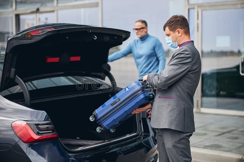 Serious elegant dark-haired Caucasian airport driver putting his client traveling bag in the car trunk. Serious elegant dark-haired Caucasian airport driver putting his client traveling bag in the car trunk