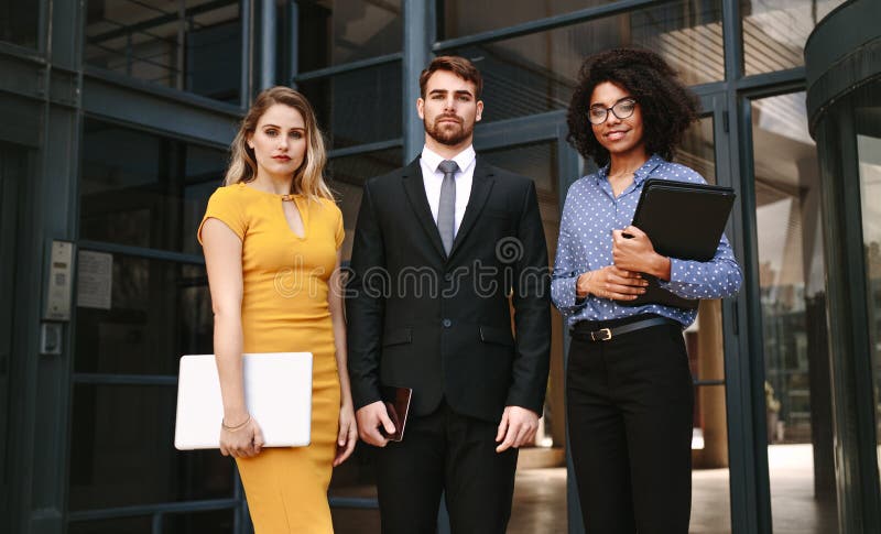 Portrait of three business people standing together in front of office building. Group of diverse business professionals looking at camera confidently. Portrait of three business people standing together in front of office building. Group of diverse business professionals looking at camera confidently.