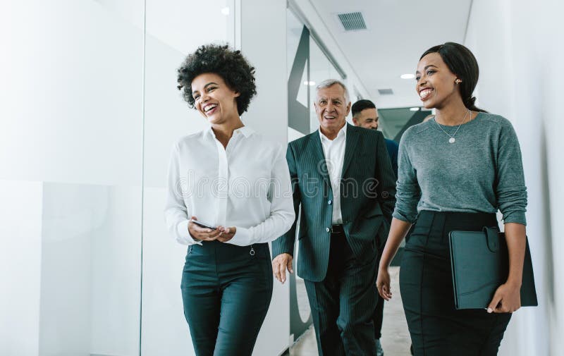 Diverse group of business people walking through office corridor. Team of corporate professionals walking and talking in modern office hallway. Diverse group of business people walking through office corridor. Team of corporate professionals walking and talking in modern office hallway.