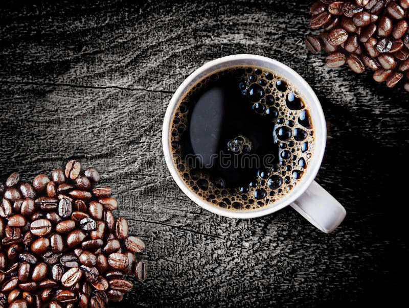 Full roast coffee beans piled on a rough rustic wooden surface with a cup of freshly brewed espresso coffee with frothy bubbles, close up overhead view. Full roast coffee beans piled on a rough rustic wooden surface with a cup of freshly brewed espresso coffee with frothy bubbles, close up overhead view