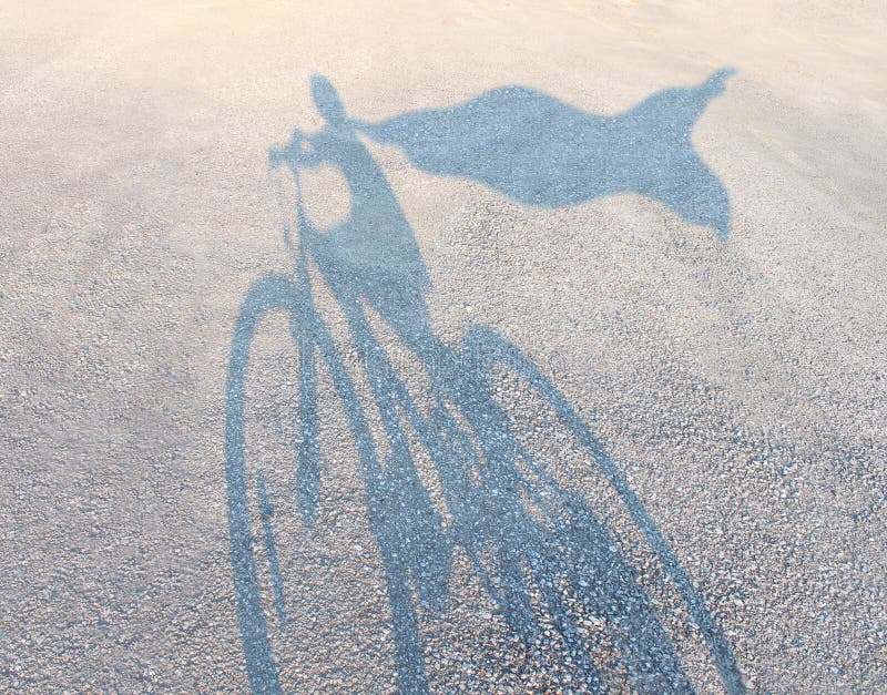 Superhero child wearing a cape riding a bicycle as a cast shadow on a road pretending to be a powerful hero as a metaphor and symbol of youth and childhood imagination and empowering kids self esteem. Superhero child wearing a cape riding a bicycle as a cast shadow on a road pretending to be a powerful hero as a metaphor and symbol of youth and childhood imagination and empowering kids self esteem.