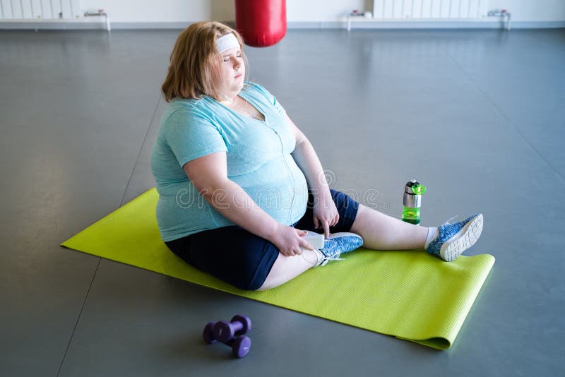 Full length portrait of tired obese woman sitting on yoga mat and resting after fitness training in gym. Full length portrait of tired obese woman sitting on yoga mat and resting after fitness training in gym