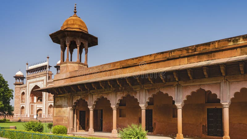 Stone gallery in the ancient Taj Mahal complex against the blue sky. Columns, openwork arches, the dome of the tower against the blue sky. A mosque is visible in the distance. India. Agra. Stone gallery in the ancient Taj Mahal complex against the blue sky. Columns, openwork arches, the dome of the tower against the blue sky. A mosque is visible in the distance. India. Agra