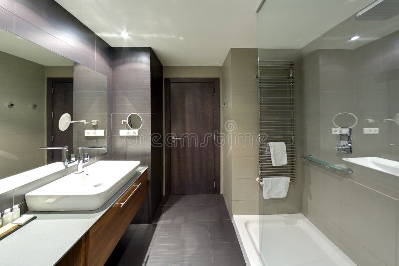 An image of a modern upscale bathroom interior in a new luxurious resort hotel. Fitted with shower area, large mirror on the wall, shaving mirror, grey marbletop sink and faucets area. Brown tile flooring. Warm lighting. Horizontal color photo format, taken with wide angle lens, nobody in picture. An image of a modern upscale bathroom interior in a new luxurious resort hotel. Fitted with shower area, large mirror on the wall, shaving mirror, grey marbletop sink and faucets area. Brown tile flooring. Warm lighting. Horizontal color photo format, taken with wide angle lens, nobody in picture.