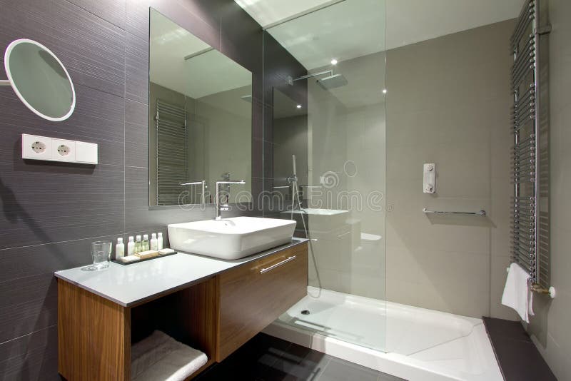 An image of a modern upscale bathroom interior in a new luxurious resort hotel. Fitted with shower area, large mirror on the wall, shaving mirror, grey marbletop sink and faucets area. Brown tile flooring. Warm lighting. Horizontal color photo format, taken with wide angle lens, nobody in picture. An image of a modern upscale bathroom interior in a new luxurious resort hotel. Fitted with shower area, large mirror on the wall, shaving mirror, grey marbletop sink and faucets area. Brown tile flooring. Warm lighting. Horizontal color photo format, taken with wide angle lens, nobody in picture.