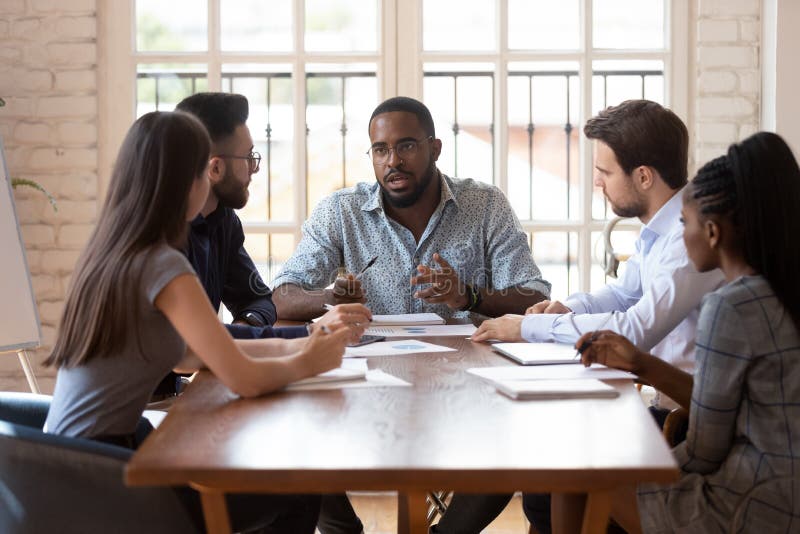 Diverse group of professionals sitting at table at brainstorming meeting, listening to african american confident male team leader, explaining planning company marketing developing strategy at office. Diverse group of professionals sitting at table at brainstorming meeting, listening to african american confident male team leader, explaining planning company marketing developing strategy at office.