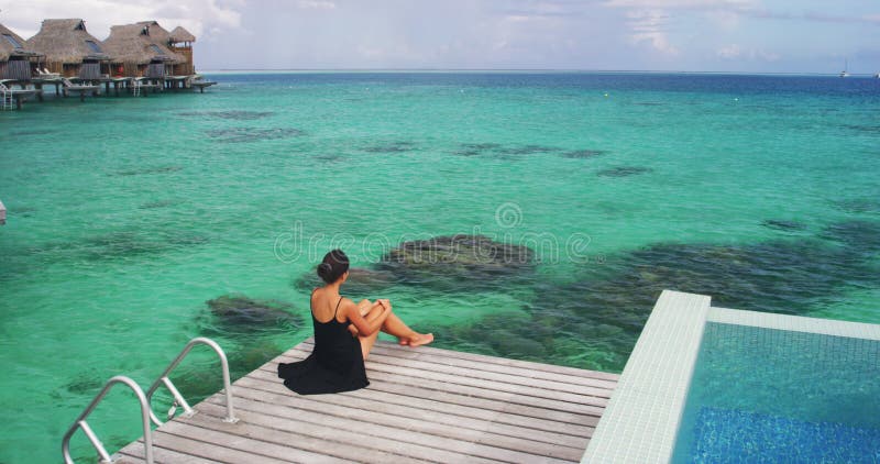 πολυτελές ταξίδι διακοπές στο bora bora. κομψή γυναίκα με μαύρο φόρεμα χαλαρώνει απολαμβάνοντας την θέα των κοραλλιογενών υφάλων σ