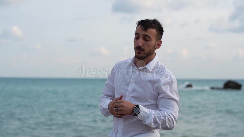 Πορτρέτο ενός στοχαστικού επιτυχούς γενειοφόρου νεαρού άνδρα που στέκεται στην παραλία και που εξετάζει το ρολόι του Νέες στάσεις