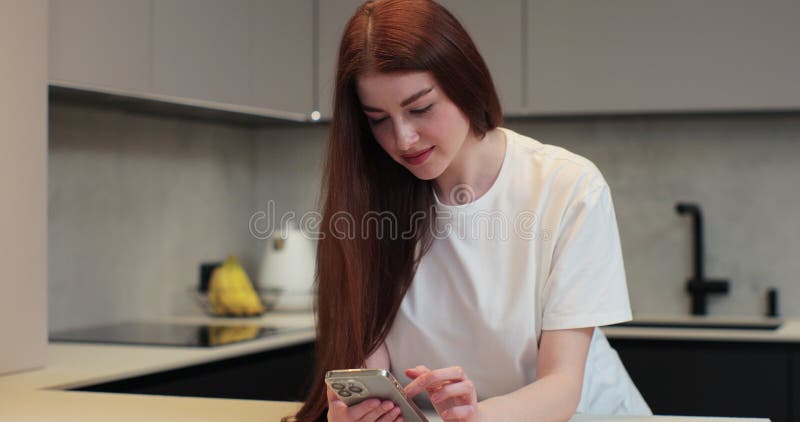 Portrait of happy young woman using smartphone in the home kitchen. Woman choosing recipe on mobile phone. Lifestyle and gadgets concept. Portrait of happy young woman using smartphone in the home kitchen. Woman choosing recipe on mobile phone. Lifestyle and gadgets concept