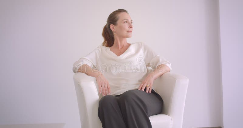 Πορτρέτο κινηματογραφήσεων σε πρώτο πλάνο της ηλικιωμένης καυκάσιας θηλυκής συνεδρίασης brunette στην πολυθρόνα σε ένα άσπρο διαμ