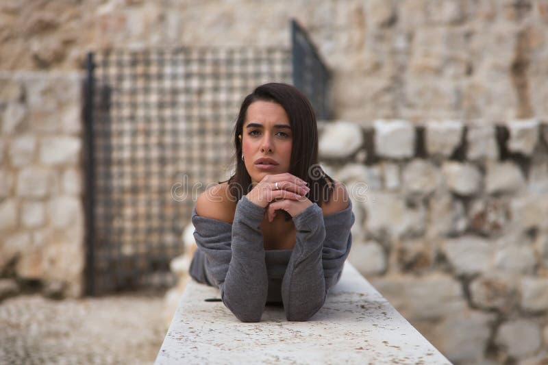 Πορτρέτο μιας νεαρής όμορφης μελαχρινής γυναίκας με γκρι φόρεμα λυπημένη και τρυφερή με τα χέρια της στο πηγούνι ξαπλωμένη πάνω σε στοκ εικόνες