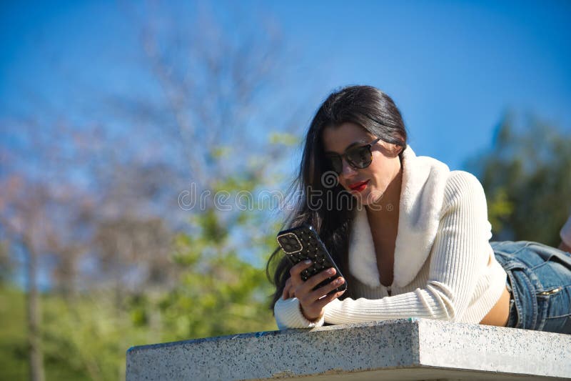 Portrait of young, beautiful, dark-haired woman wearing a white sweater and sunglasses, checking social media on her smartphone lying on a park bench on a sunny day. Beauty, fashion, trend concept. Portrait of young, beautiful, dark-haired woman wearing a white sweater and sunglasses, checking social media on her smartphone lying on a park bench on a sunny day. Beauty, fashion, trend concept