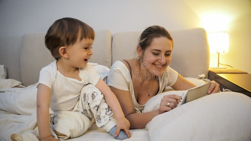 Πορτρέτο της χαμογελώντας νέας μητέρας με το γιο μικρών παιδιών που χρησιμοποιεί τον ψηφιακό υπολογιστή ταμπλετών στο κρεβάτι στοκ φωτογραφία με δικαίωμα ελεύθερης χρήσης Πορτρέτο της χαμογελώντας νέας μητέρας με το γιο μικρών παιδιών που χρησιμοποιεί τον ψηφιακό υπολογιστή ταμπλετών στο κρεβάτι στοκ φωτογραφία με δικαίωμα ελεύθερης χρήσης