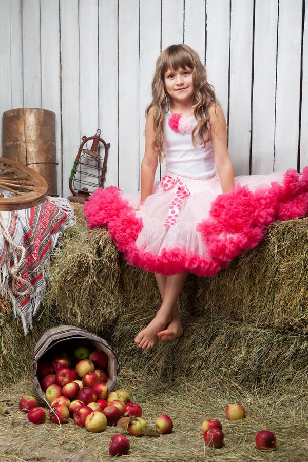 Portrait of barefooted funny little blond girl, villager dressed luxuriant wavy ball dress with frill sitting on haystack near inverted pail with apples in wooden vintage hayloft. Portrait of barefooted funny little blond girl, villager dressed luxuriant wavy ball dress with frill sitting on haystack near inverted pail with apples in wooden vintage hayloft