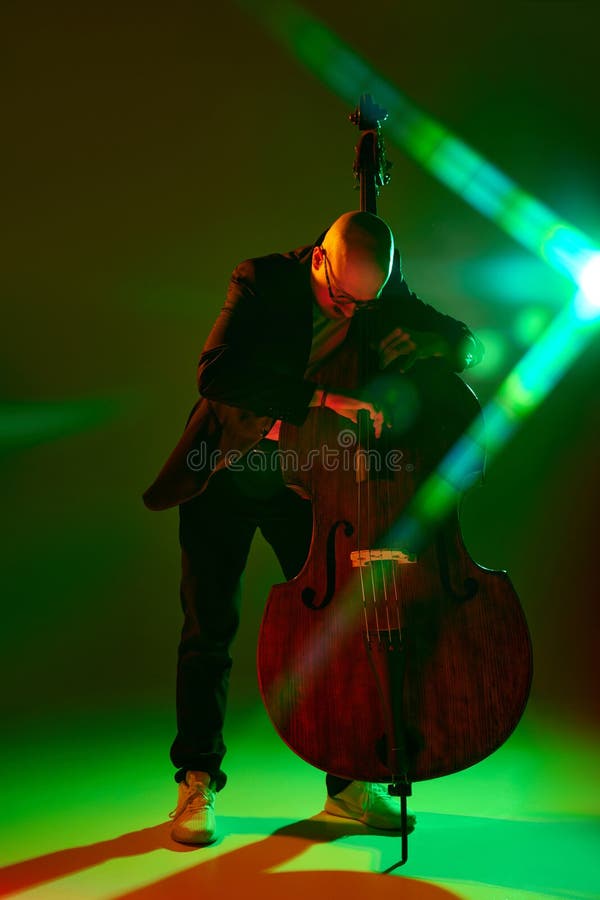 Portrait of bald musician in glasses performing on double bass in red-green neon light against gradient studio background. Concept of music and art, hobby, concerts and festivals, modern culture. Ad. Portrait of bald musician in glasses performing on double bass in red-green neon light against gradient studio background. Concept of music and art, hobby, concerts and festivals, modern culture. Ad